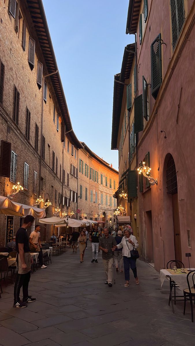 people are walking through an alleyway in the evening time, with tables and umbrellas