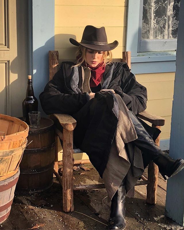 a woman sitting on a wooden chair next to a barrel and a bottle in front of a house