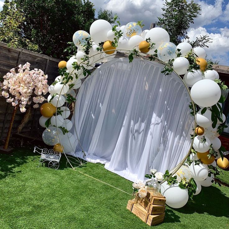 a white and gold wedding arch decorated with balloons, flowers and greenery for an outdoor ceremony