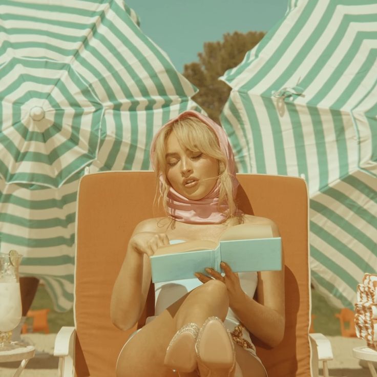 a woman sitting in a chair reading a book on the beach with umbrellas behind her