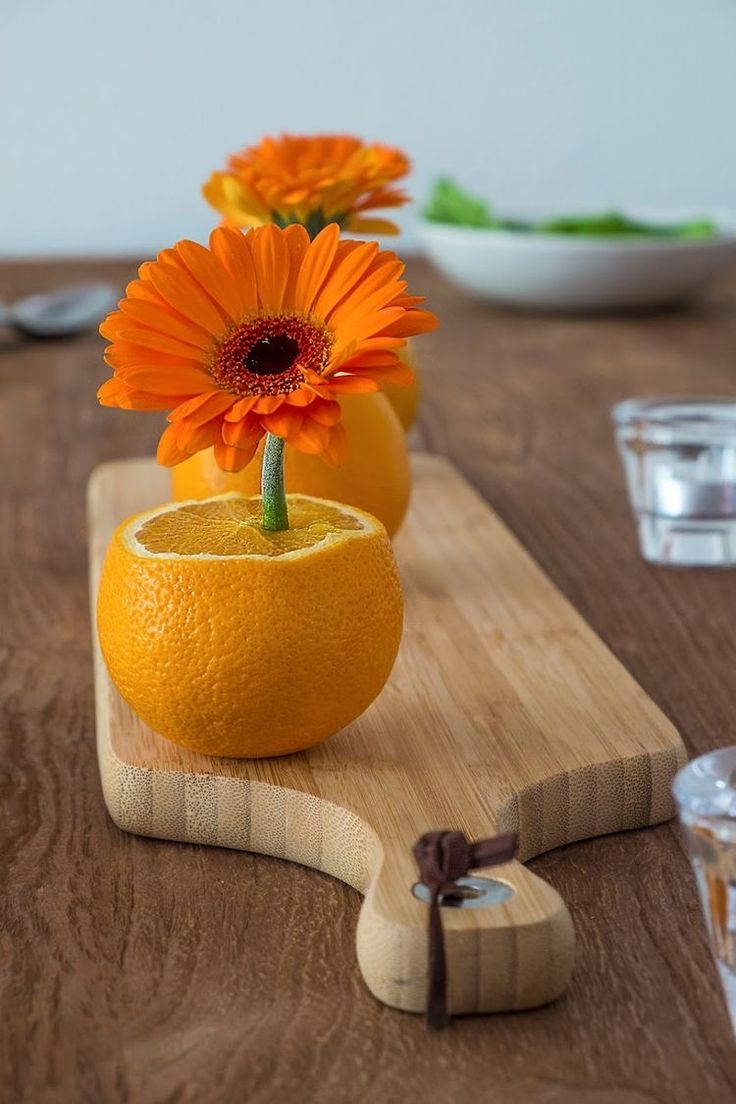 an orange sitting on top of a wooden cutting board next to two vases filled with flowers