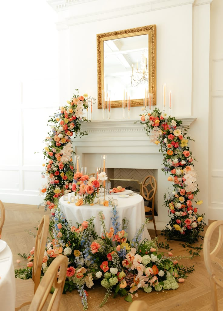 a table with flowers and candles on it in front of a fire place surrounded by chairs