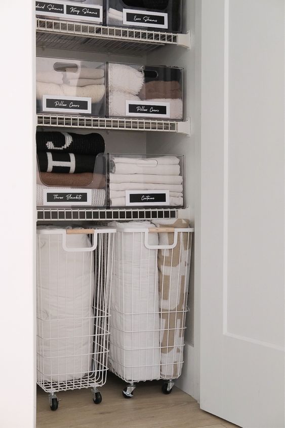 the closet is organized with baskets and linens in white bins on casteors