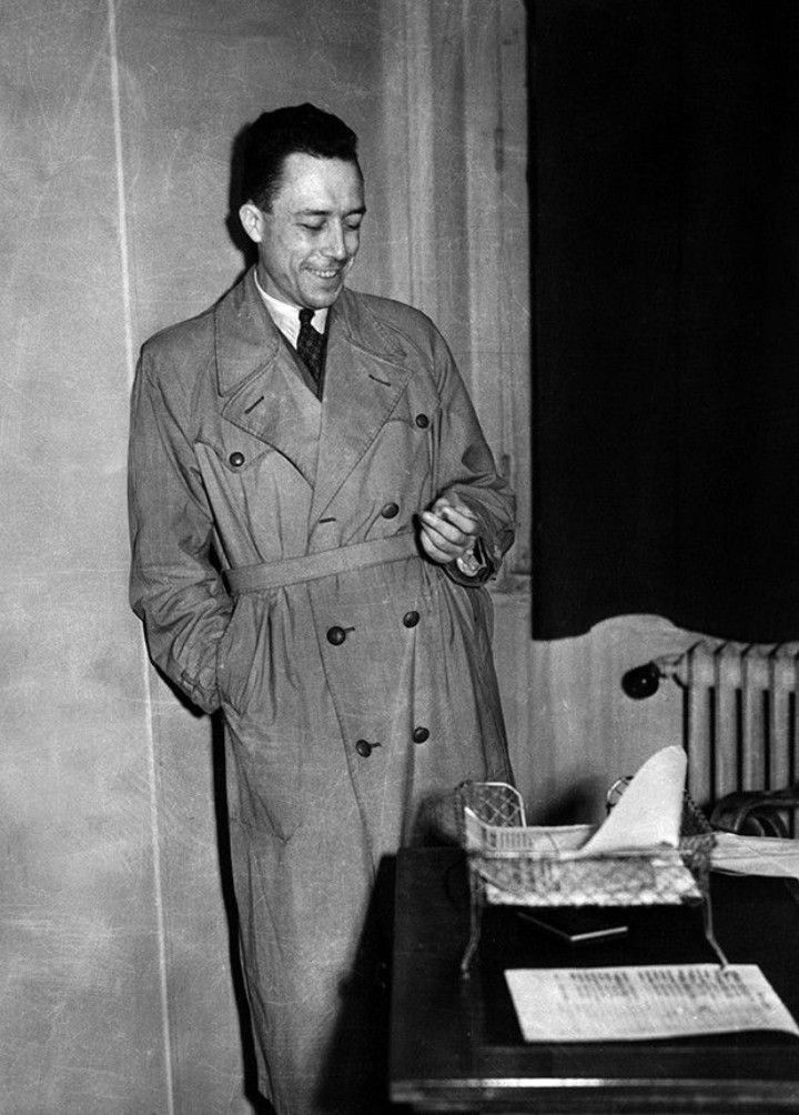 a black and white photo of a man in a trench coat standing next to a desk