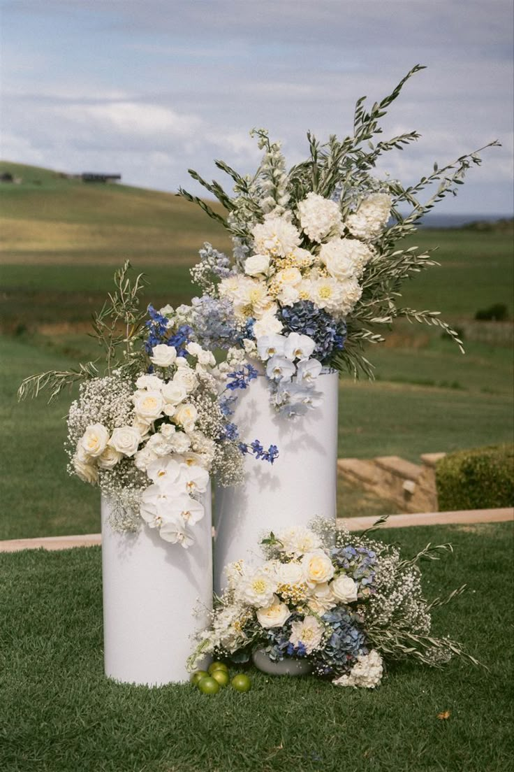 three tall white vases with flowers and greenery on the grass in front of an open field