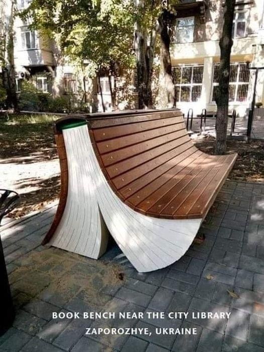 a curved wooden bench sitting on top of a brick walkway next to a tree filled park