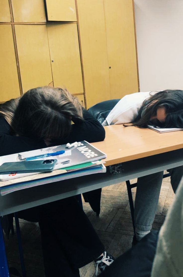 two people sitting at a table with their heads on the desk and one person sleeping