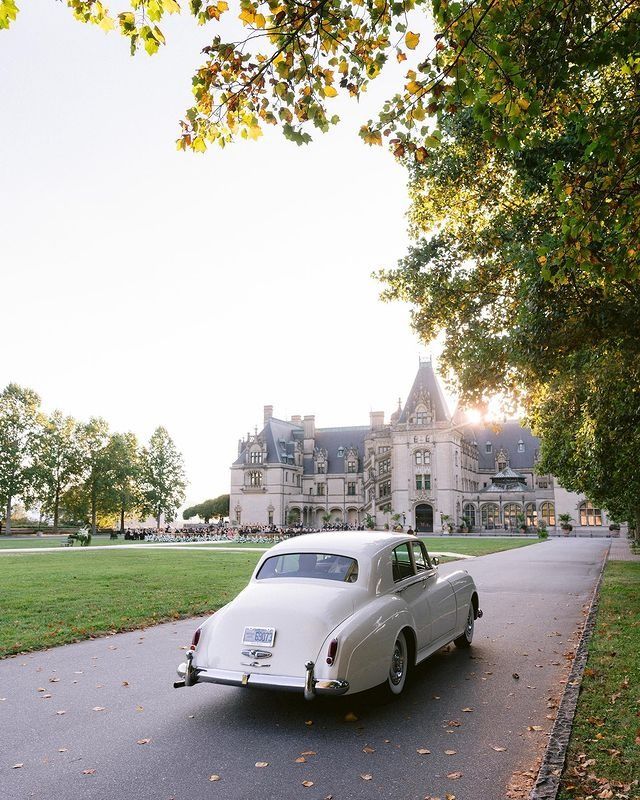 an old white car is parked in front of a large house on the side of a road