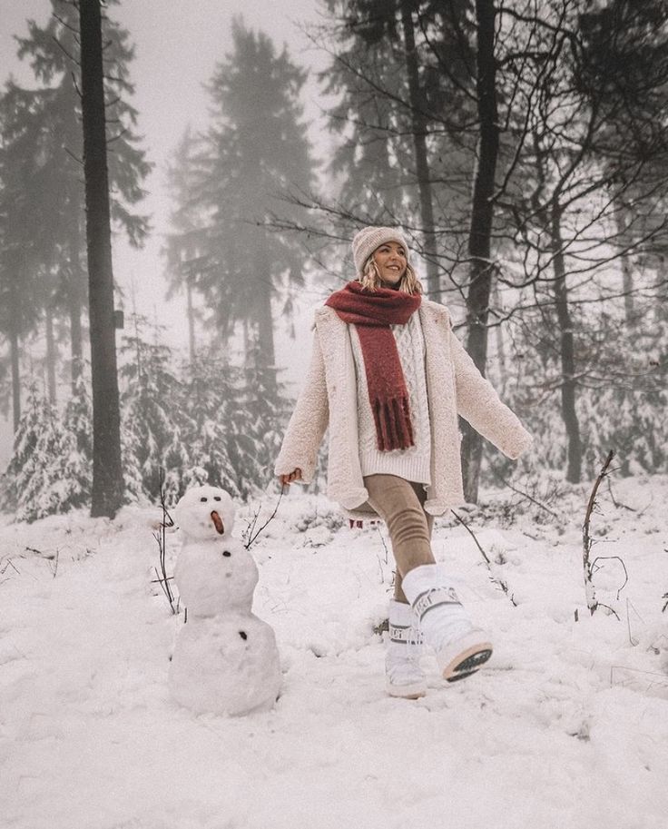 a woman standing next to a snowman on top of a snow covered ground with trees in the background