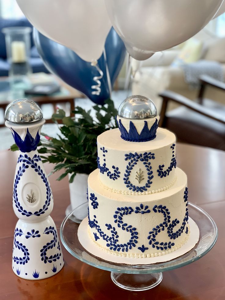 a blue and white cake on a glass plate with balloons in the air behind it