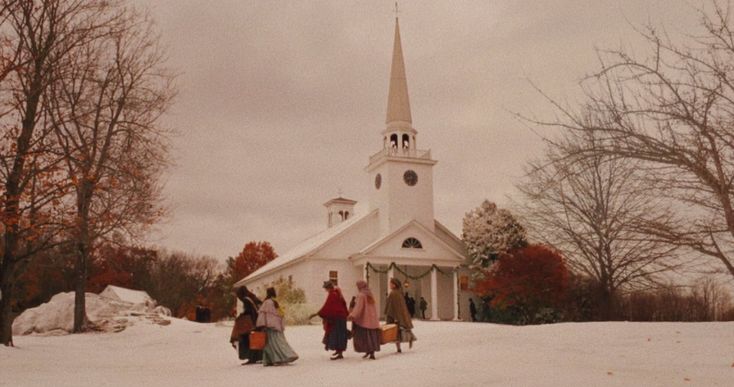 a group of people standing in front of a white church with a steeple covered in snow