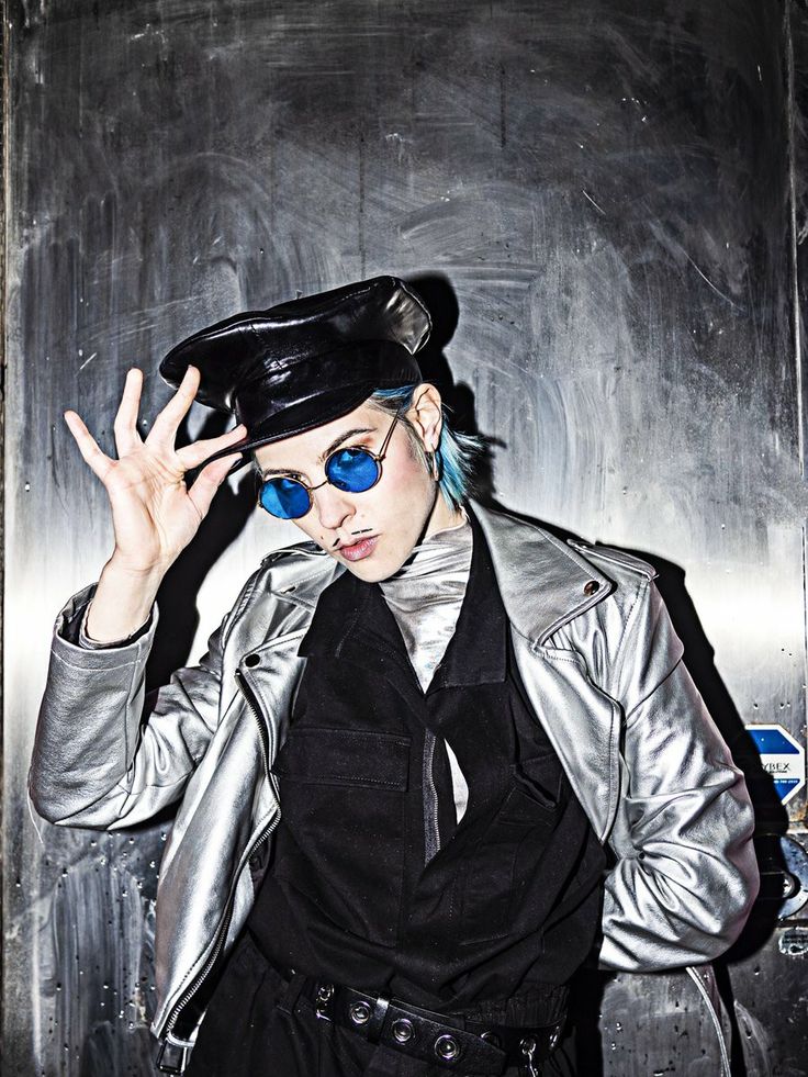 a woman wearing sunglasses and a hat is standing in front of a metal wall with her hand on her head