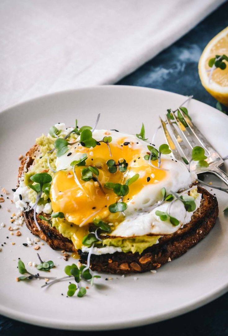 an open face sandwich topped with eggs and green onions on a white plate next to a lemon slice