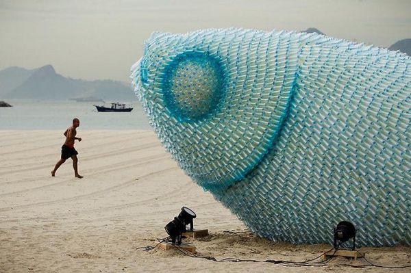 a man running on the beach next to a giant fish sculpture that looks like it's made out of plastic bottles