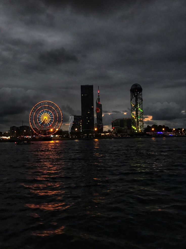 the ferris wheel is lit up at night by the water's edge with buildings in the background