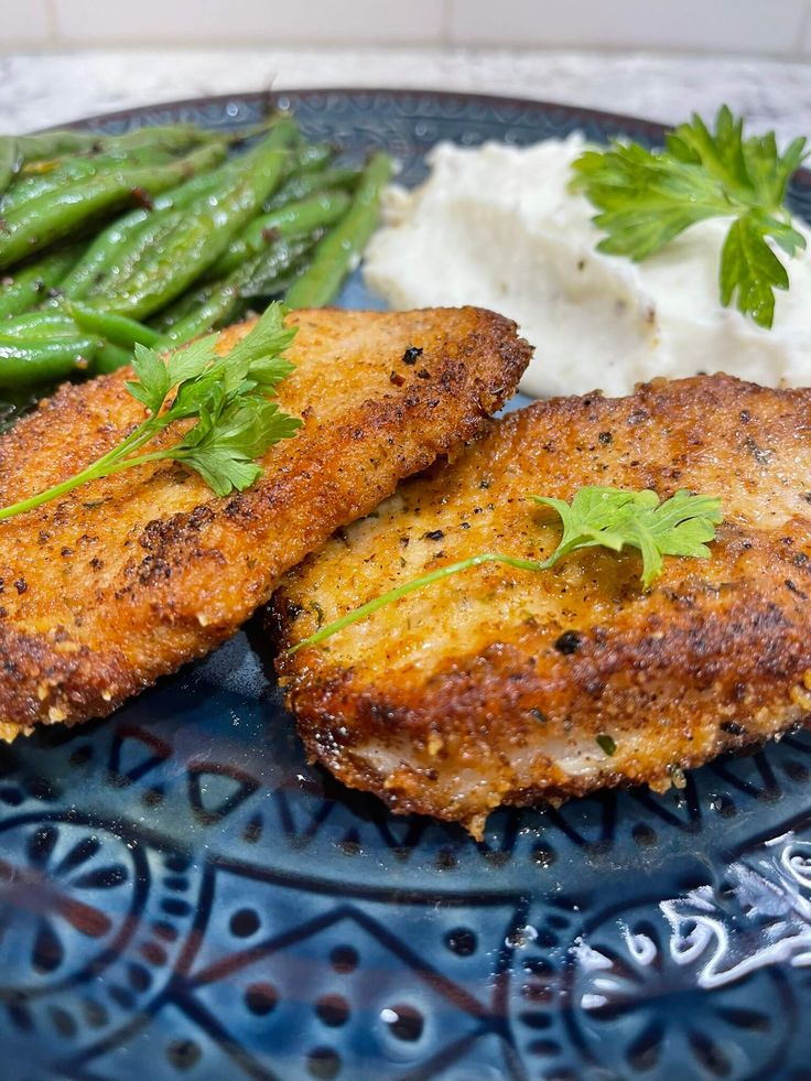 two pieces of fried chicken on a blue plate with mashed potatoes and green beans