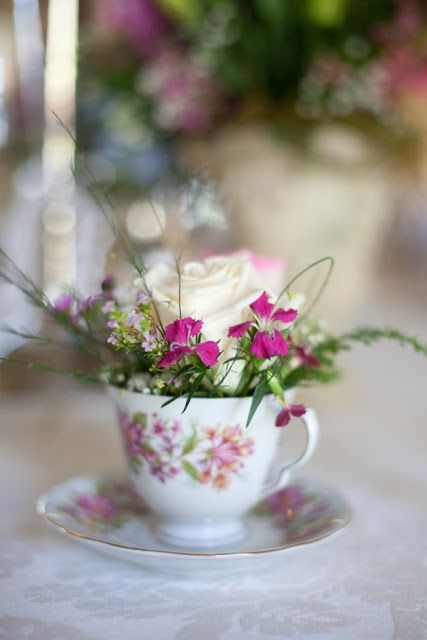 a white rose in a teacup on a table with pink and purple flowers behind it