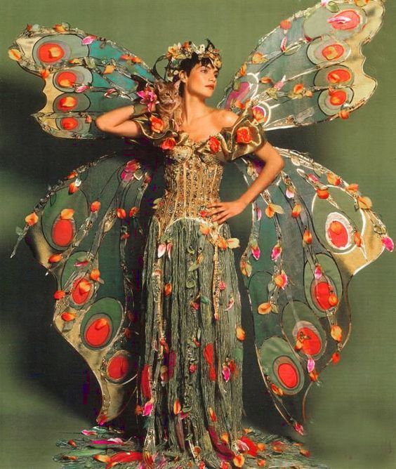 a woman dressed as a peacock with wings and flowers on her head, standing in front of a green background