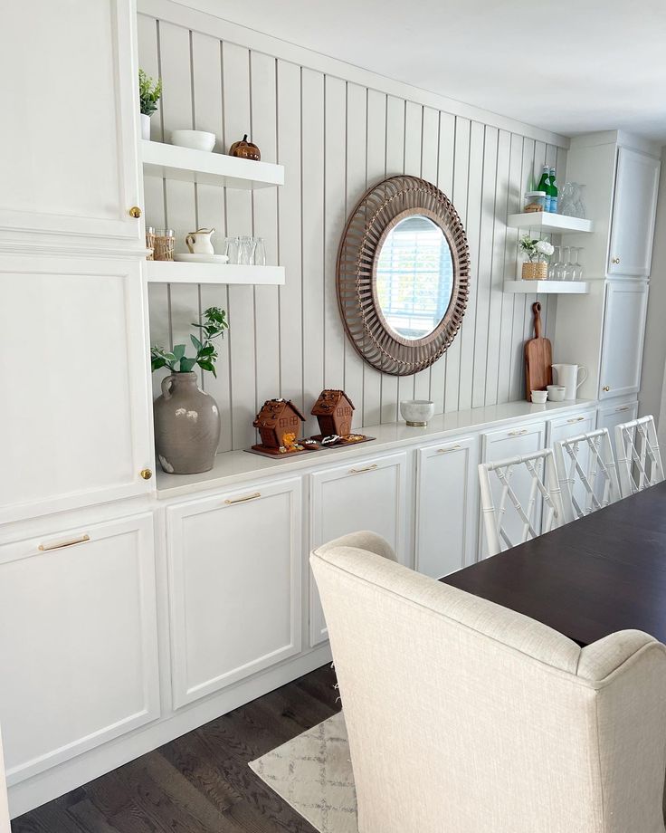 a dining room table with chairs and white cabinets in the back ground, there is a round mirror on the wall above it