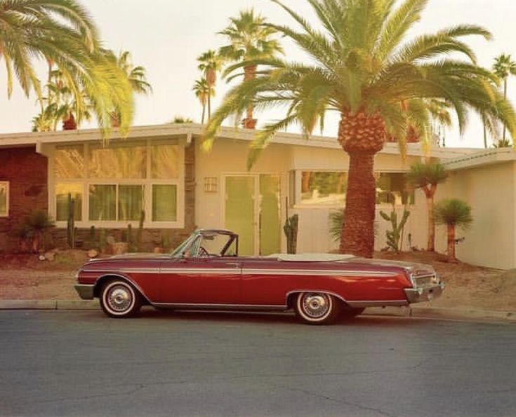 an old red car parked in front of a house with palm trees on the side