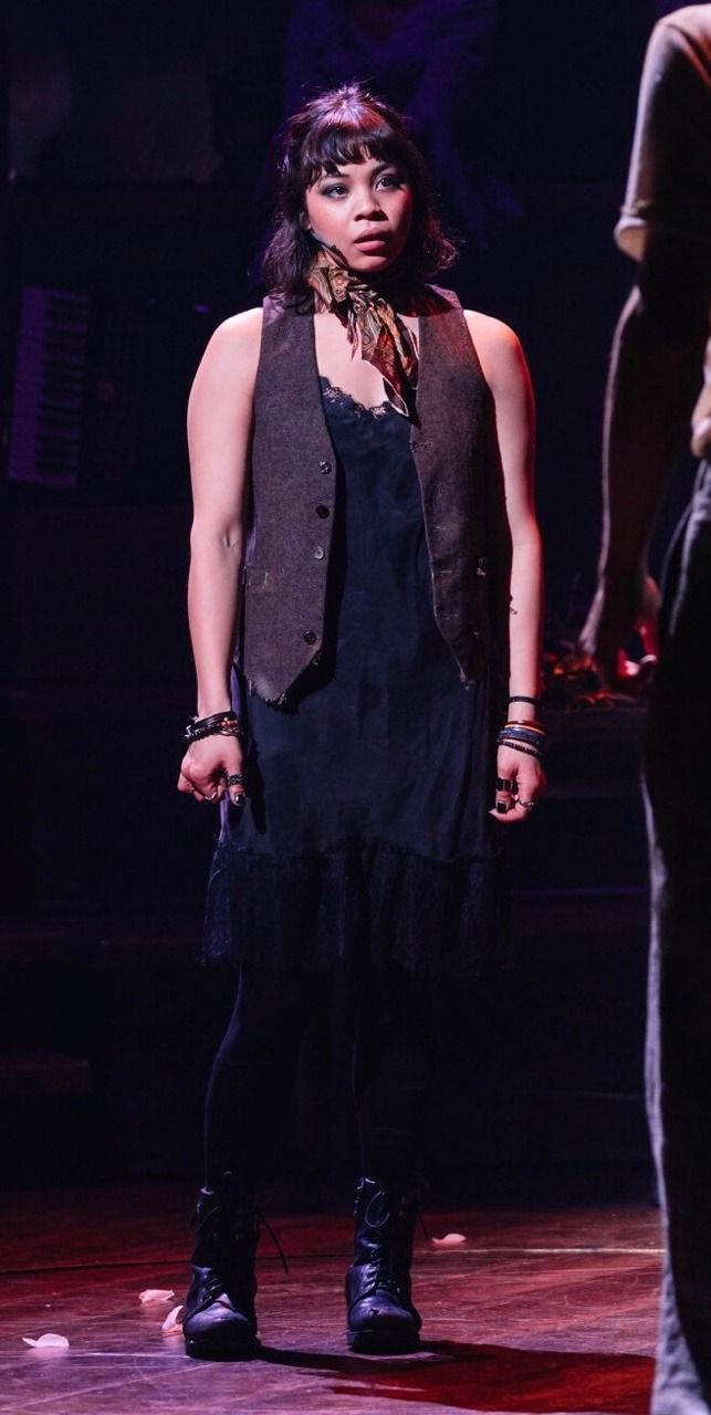 a woman is standing on stage wearing a black dress and vest with her hands in her pockets