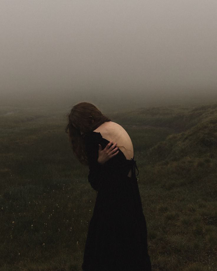 a woman in a black dress standing on a foggy field with her back to the camera