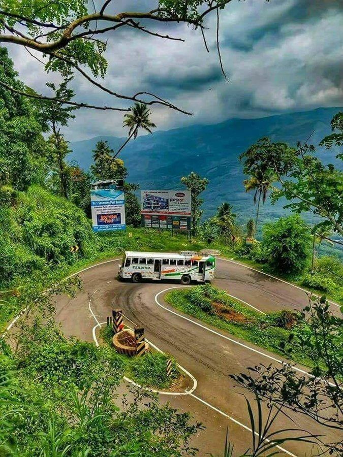 a bus driving down a road next to lush green trees and mountains on a cloudy day