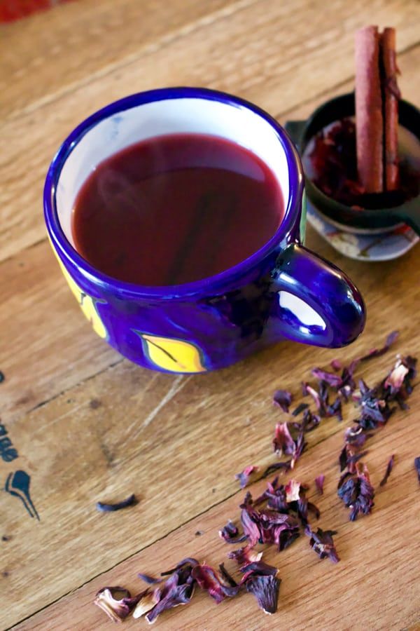 a cup of tea next to a bowl of dried rose petals and cinnamon sticks on a wooden table