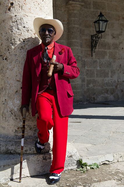 a man in a red suit and hat leaning against a stone wall with a cane