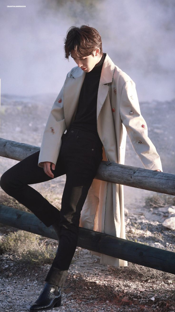 a man leaning on a fence wearing a trench coat and black pants with his hands in his pockets