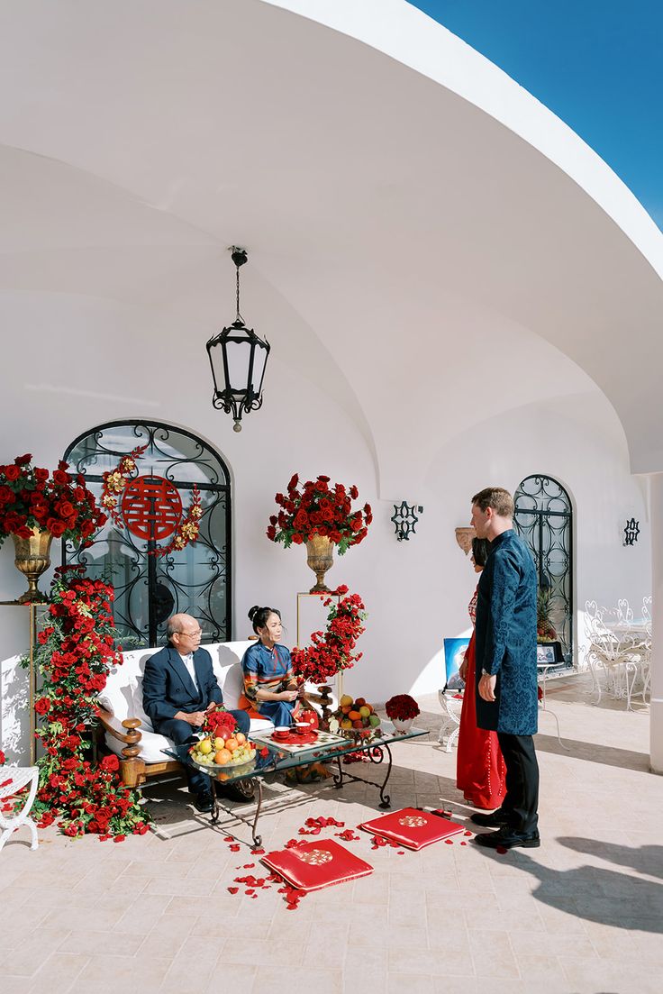 three men sitting on couches in front of a white building with red flowers and decorations