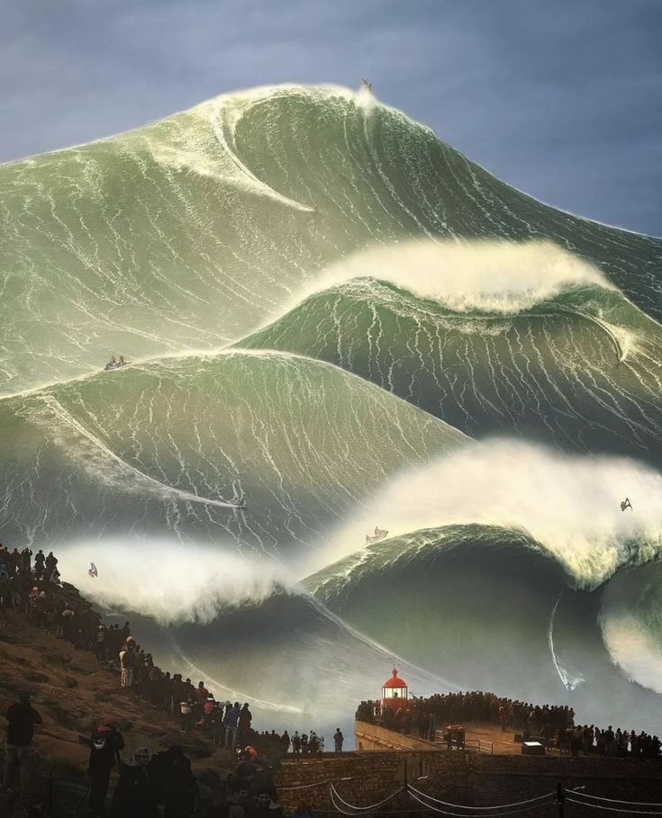 people are standing on the side of a hill watching large waves crash over them in front of a lighthouse