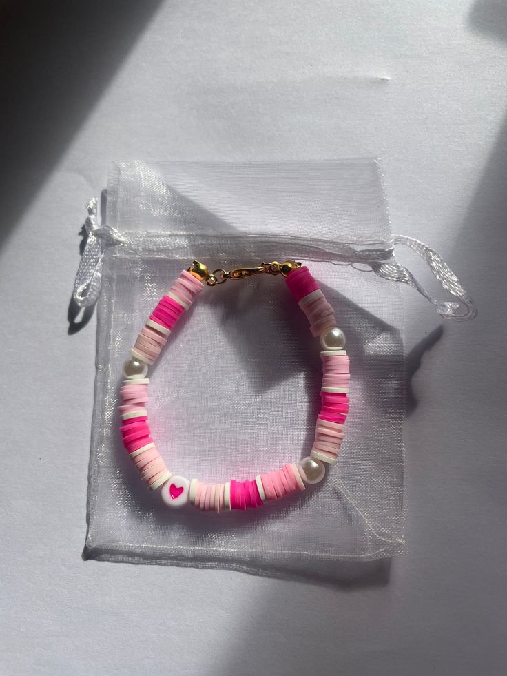 a pink and white beaded bracelet sitting on top of a clear bag with a gold charm