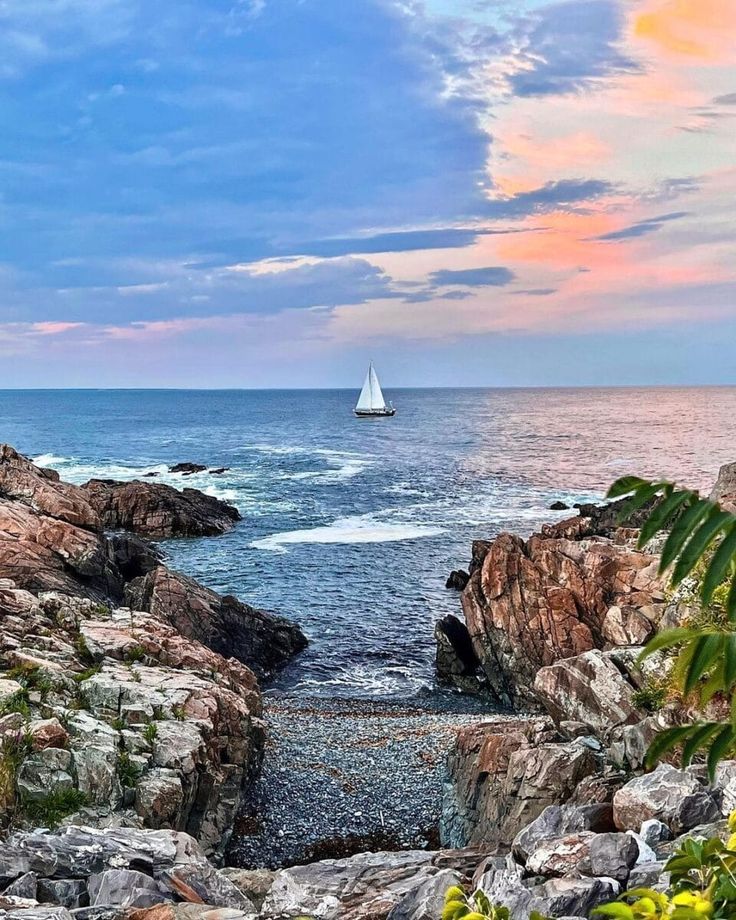 a sailboat is out on the water near some rocks and greenery at sunset