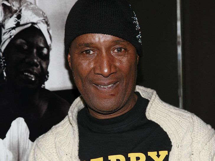 a man wearing a black beanie smiles at the camera while standing in front of a poster