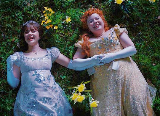 two women in dresses standing next to each other with flowers on the ground behind them