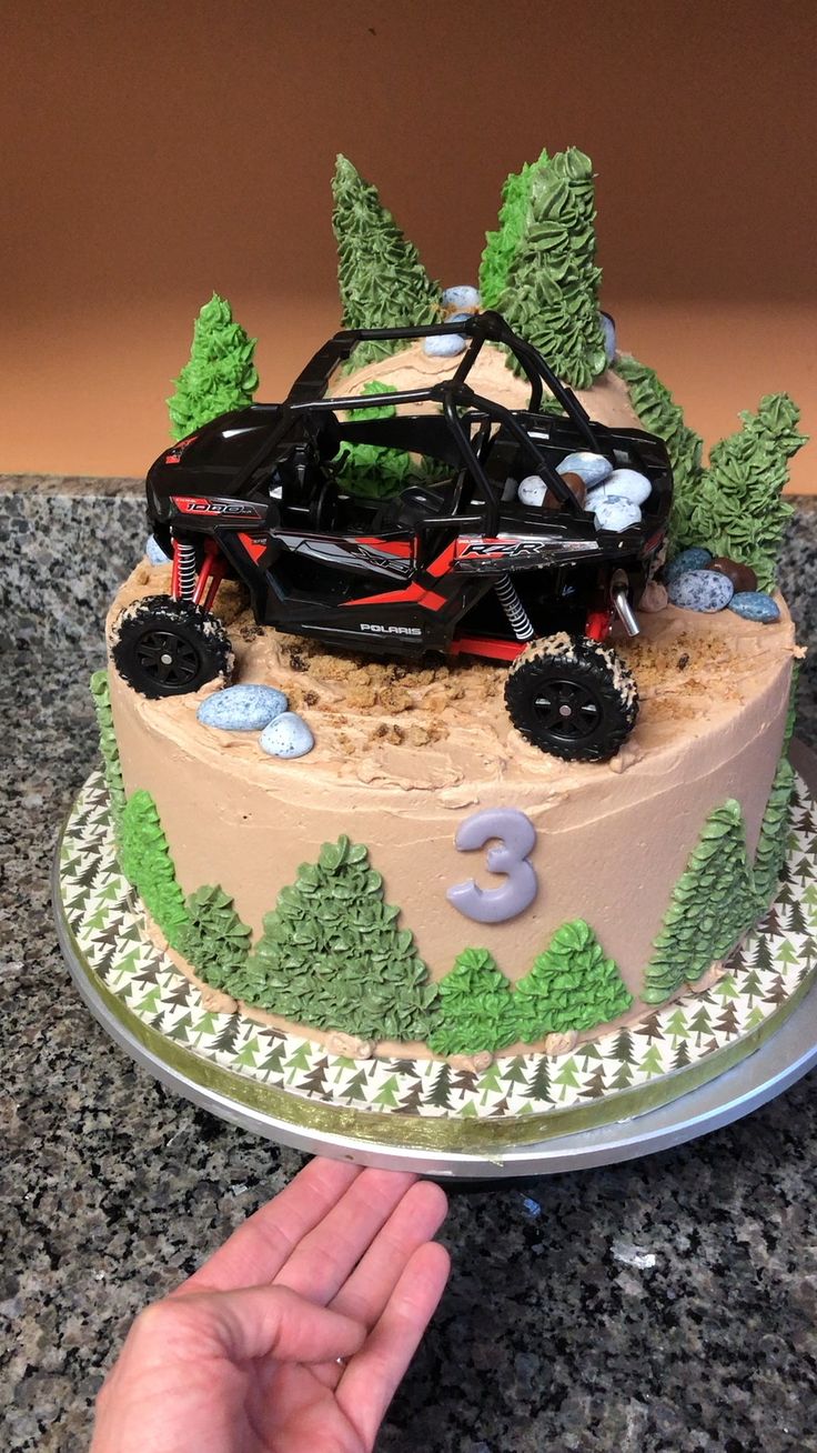 a birthday cake with a remote control car on it's top and trees in the background