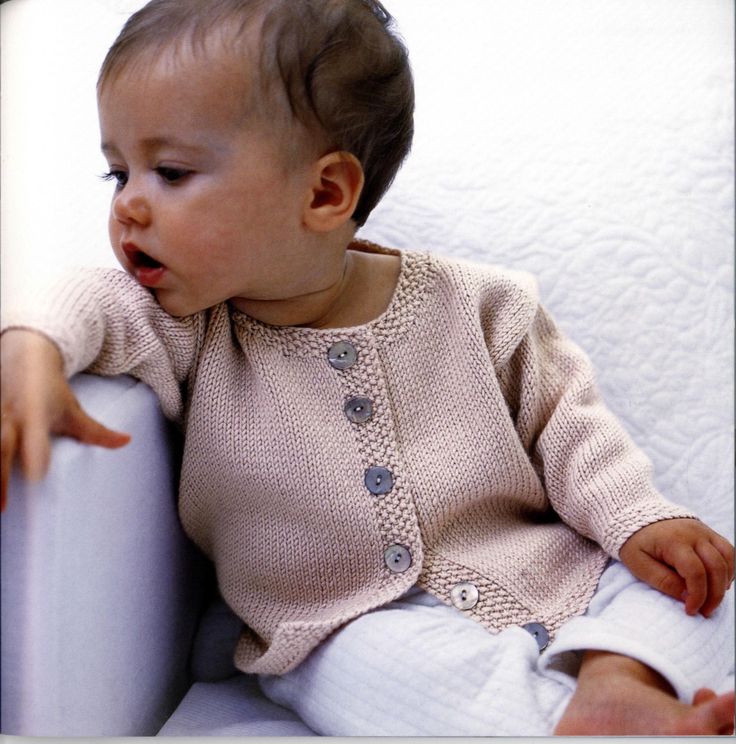 a baby sitting on a white couch wearing a knitted sweater and pants with buttons