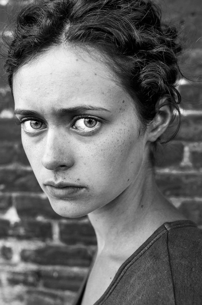 a black and white photo of a woman in front of a brick wall with her eyes wide open