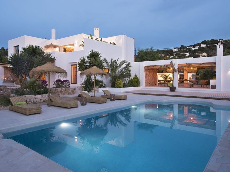 an outdoor swimming pool with lounge chairs and umbrellas next to the house at dusk