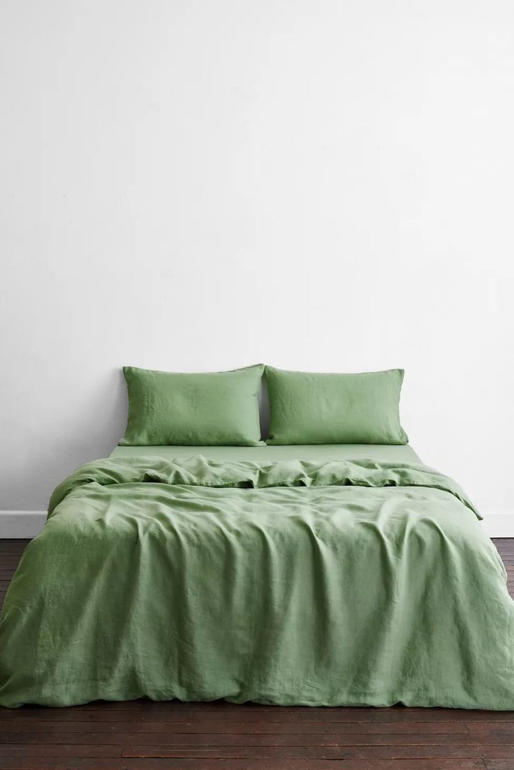 a bed with green sheets and pillows in a white room next to a wooden floor