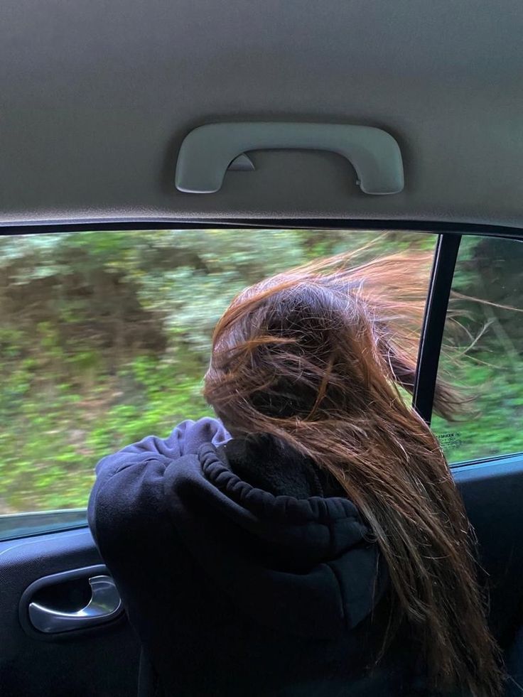 a woman sitting in the passenger seat of a car looking out the window at trees