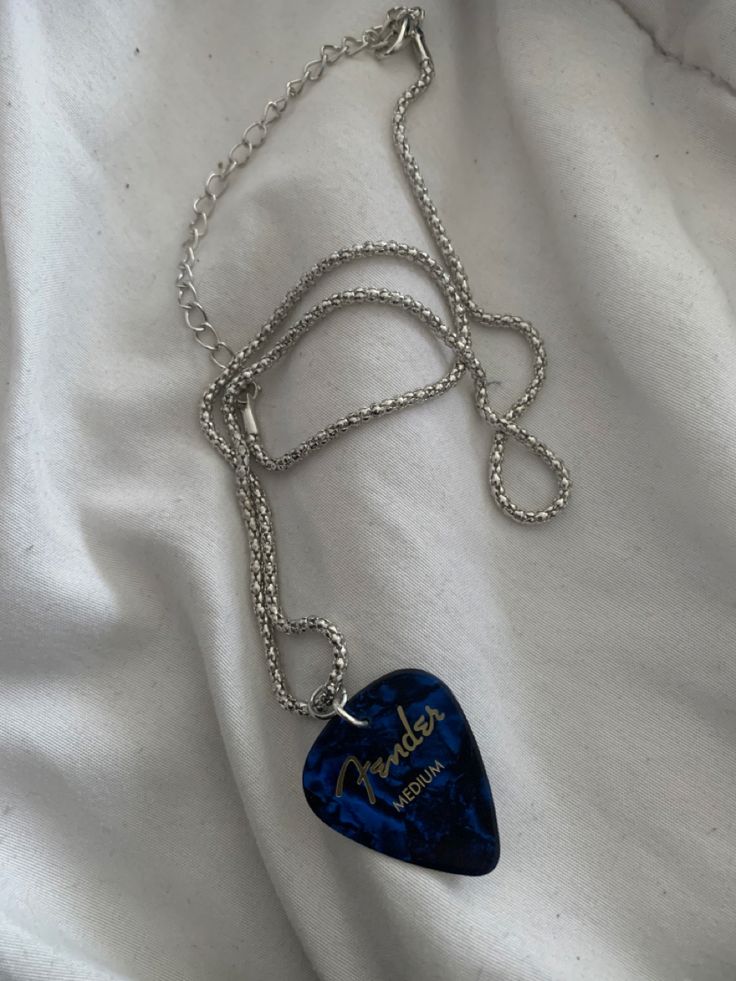 a blue guitar pick on a chain with the word fender written in gold lettering, sitting on a white sheet