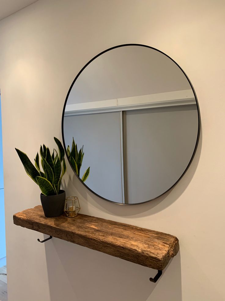 a round mirror hanging on the wall above a wooden shelf with a potted plant