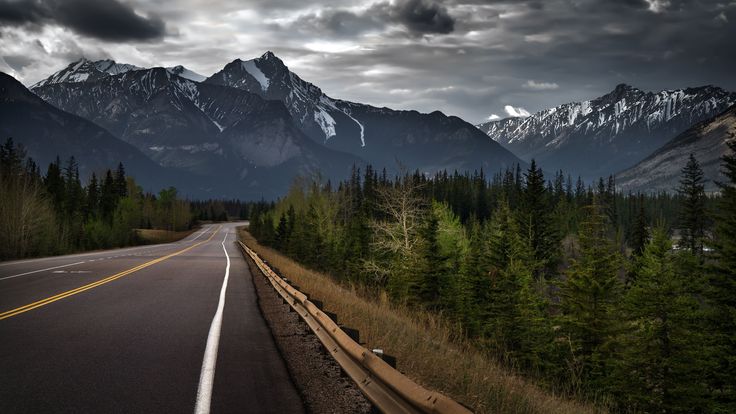 an empty road with mountains in the background and trees on both sides, under a cloudy sky