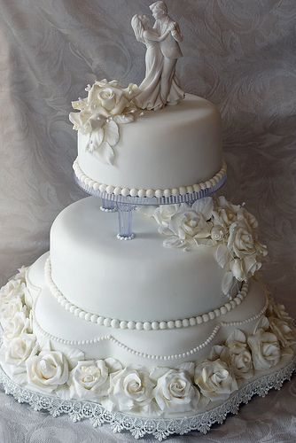 a three tiered wedding cake with white roses and pearls on the bottom is decorated with a bride and groom figurine