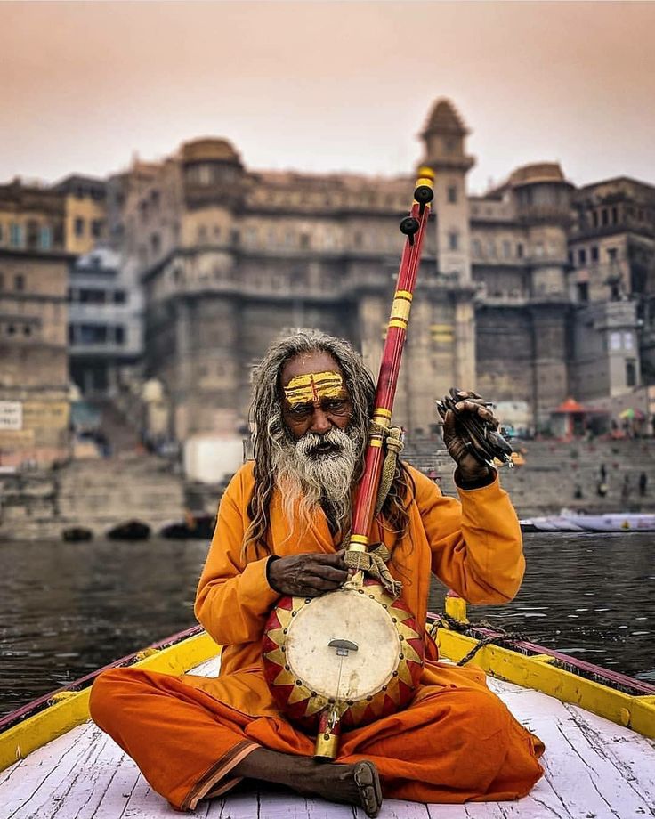 an old man sitting in a boat with a drum on the water and buildings behind him