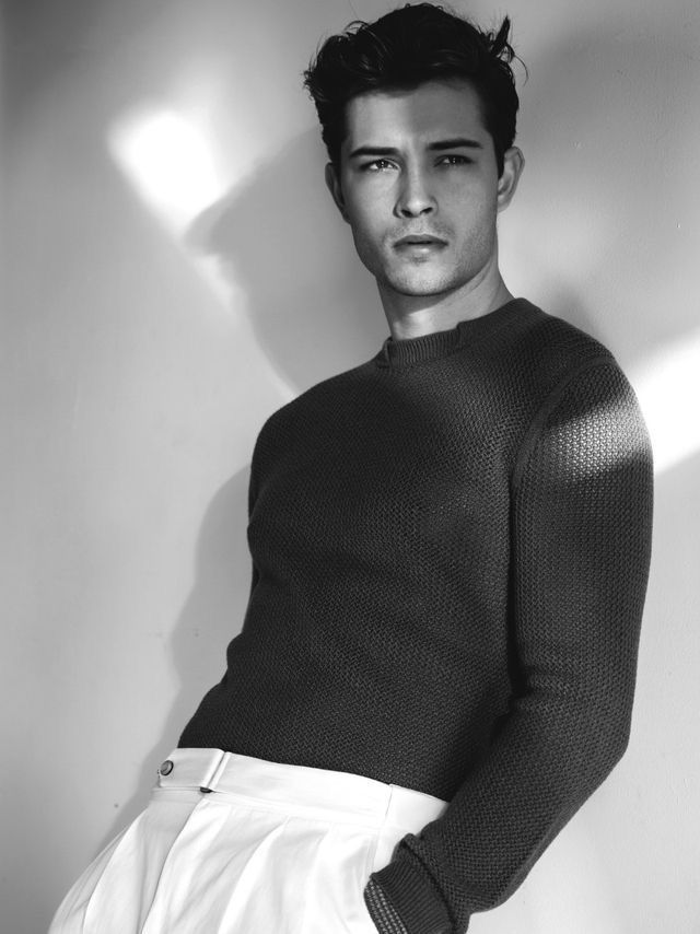 black and white photograph of a man in a sweater leaning against a wall with his hands on his hips