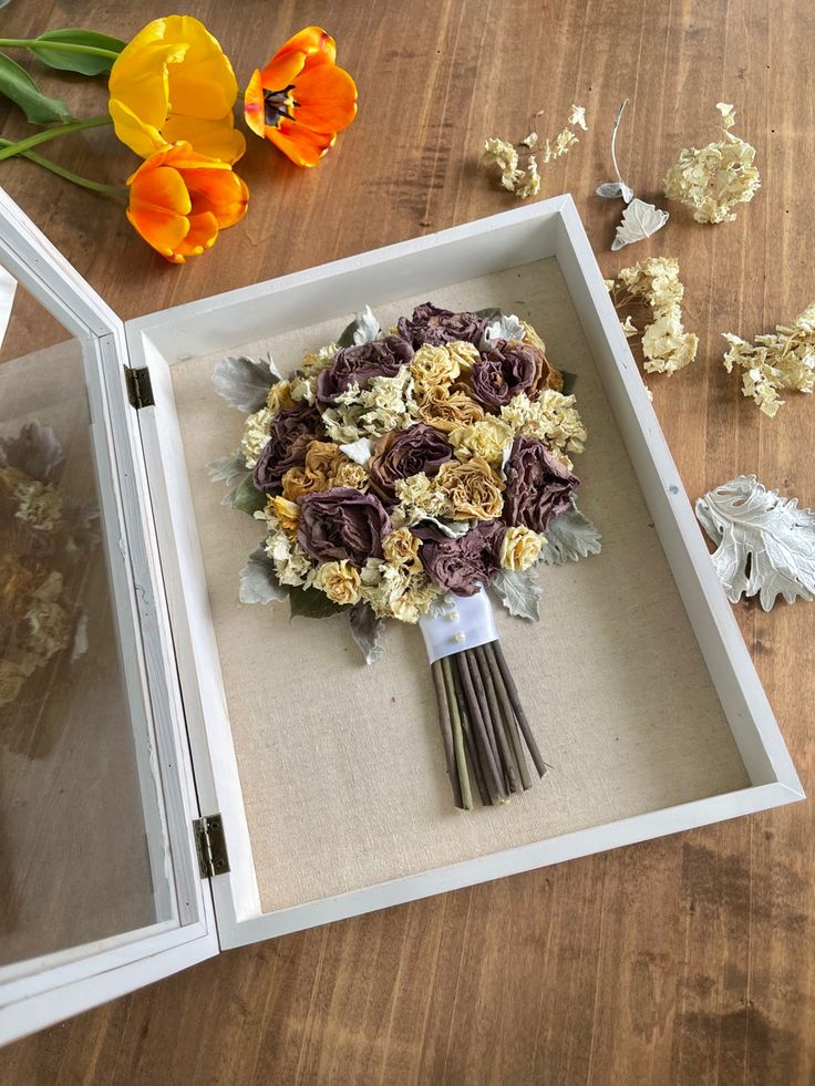 an arrangement of flowers in a white box on a wooden table next to dried flowers