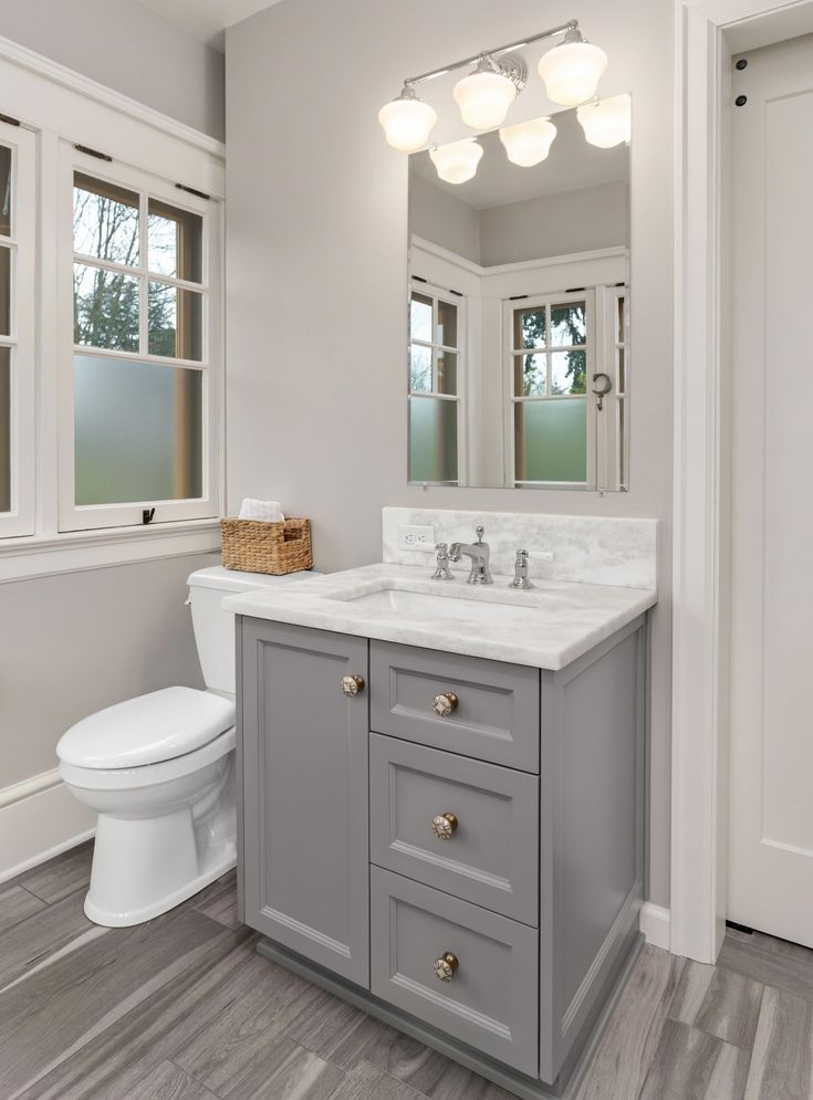 a white toilet sitting next to a sink in a bathroom under two windows on top of a hard wood floor